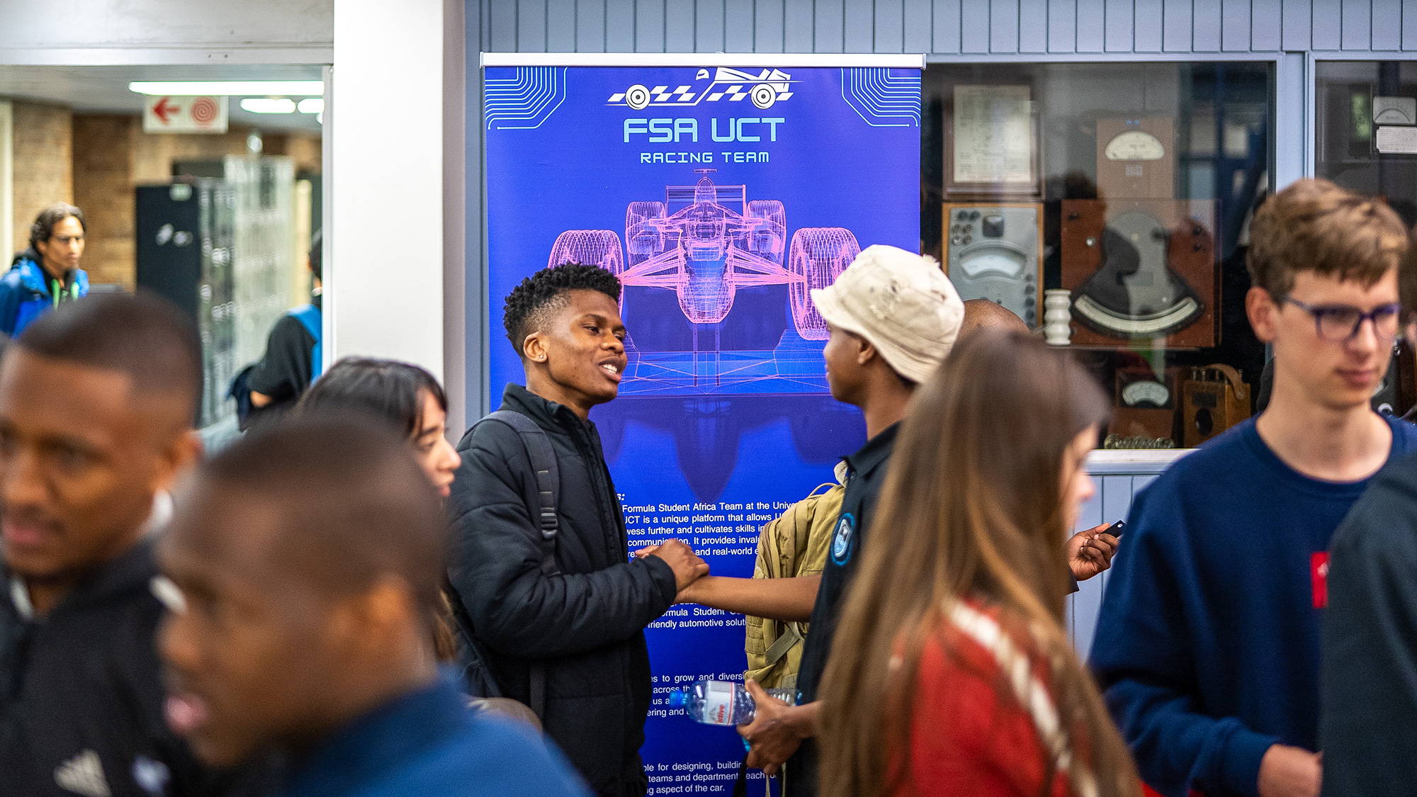 Students crowded around the UCT Racing stand at a university recruitment event, in front of a tall blue banner showing a wireframe drawing of a formula car.