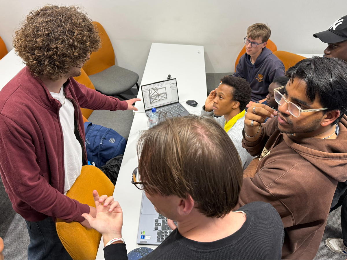 Students crowded around a laptop reviewing a wireframe chassis drawing, one pointing at the screen mid-explanation.